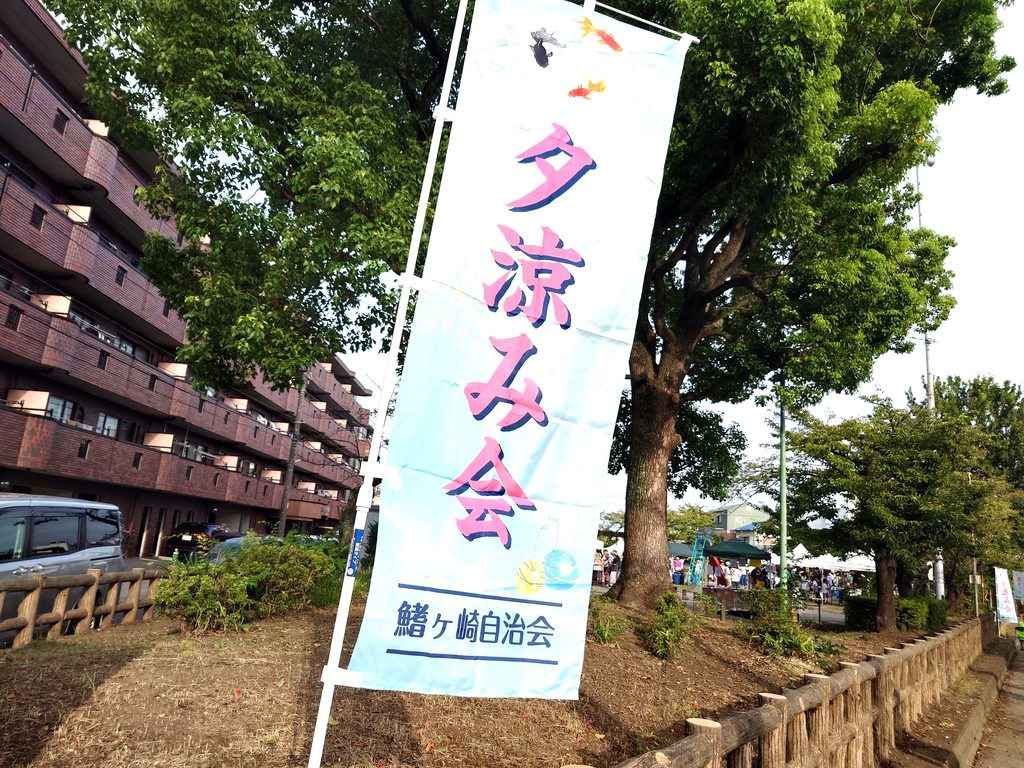 鰭ヶ崎自治会 夏まつりが開催された三角公園の様子。『夕涼み会 鰭ヶ崎自治会』と書かれたのぼりが立ち、奥には多くの来場者が集まっている。